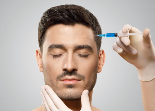 Young man isolated on gray background receiving botox injection from beauty doctor in sterile gloves in forehead to reduce aging, eyes closed