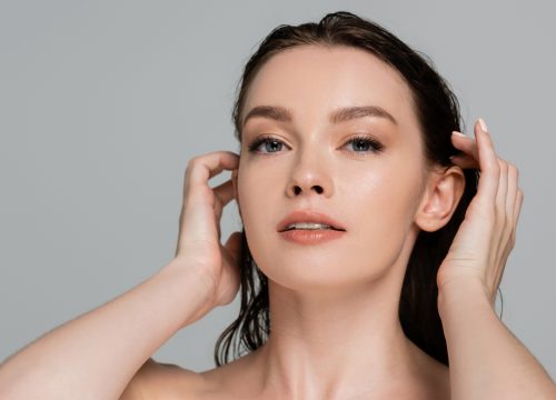 portrait of young and pretty woman adjusting wet hair while looking at camera isolated on grey