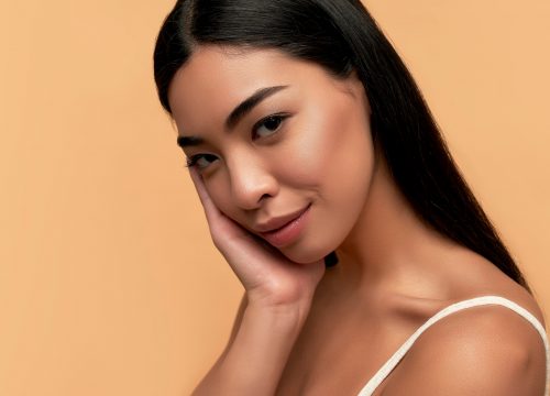 Young woman with clean healthy glowing skin in white top isolated on beige background.