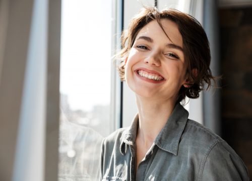 Image of beautiful young joyful woman smiling and looking at camera while standing near window
