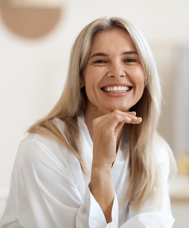 Happy attractive blonde woman in white silky bathrobe posing at white bathroom, smiling at camera