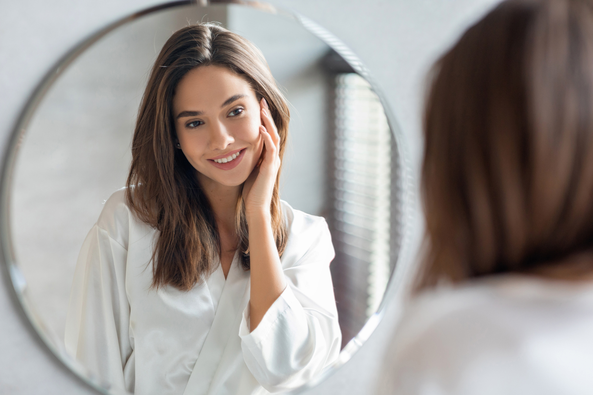 Young Attractive Woman Looking At Mirror In Bathroom after Sublime Skin Tightening treatments