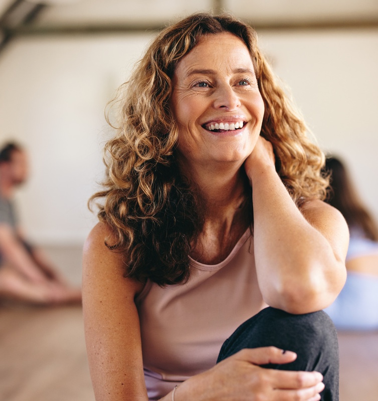 Smiling senior woman sitting in a yoga studio with a group of people exercising in the background. 